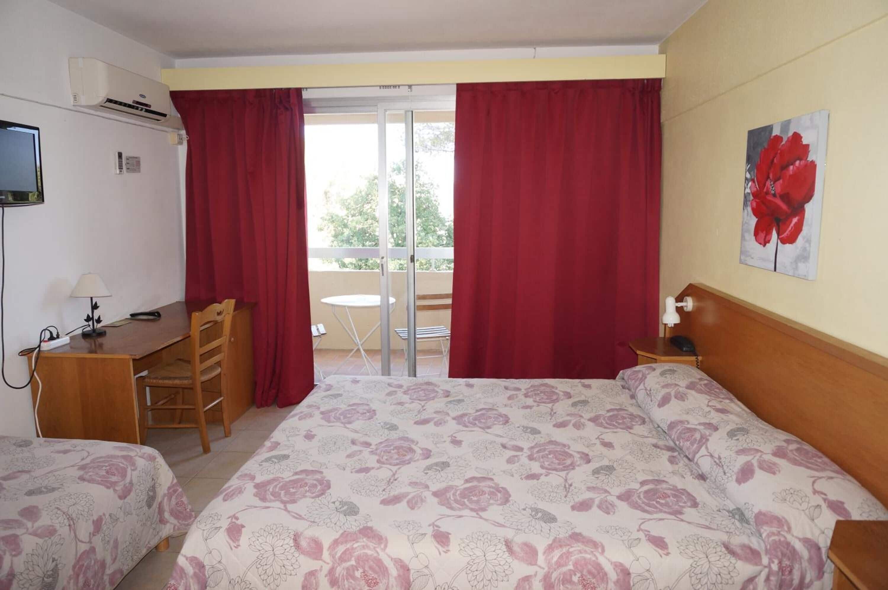 A hotel room with a bed, a desk, a chair, and a small table. There is a window with red curtains, a painting on the wall, and an air conditioning unit.