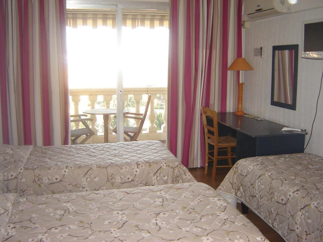 A room with two beds, a desk, and a chair. There is a balcony with a table and chairs outside the window.