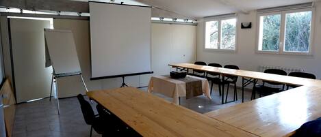 A conference room with a large table, chairs, a projector screen, and windows with a view of trees.
