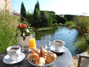 A table with croissants, coffee cups, and a glass of orange juice by a pond. - Hotel Des Grands Vins (Fleurie)