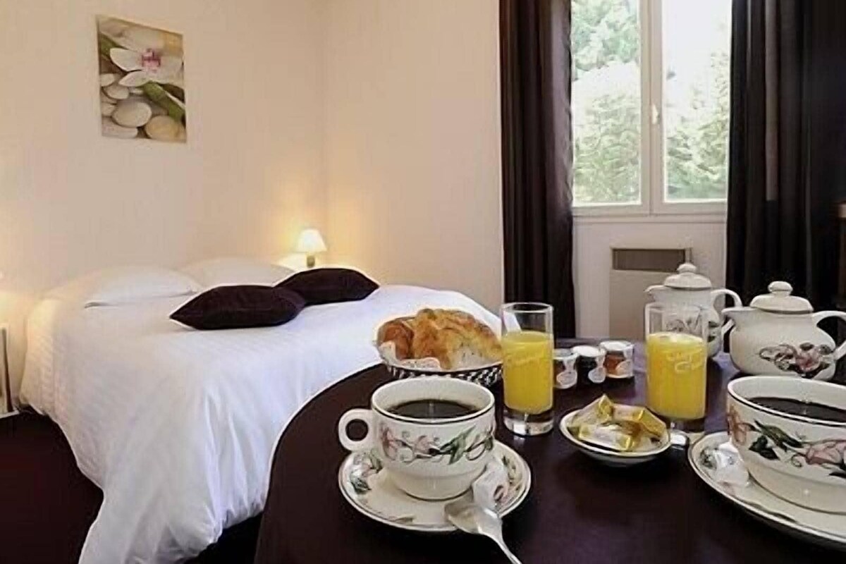 A hotel room with a neatly made bed, a breakfast table with coffee, tea, and juice, and a window with curtains.