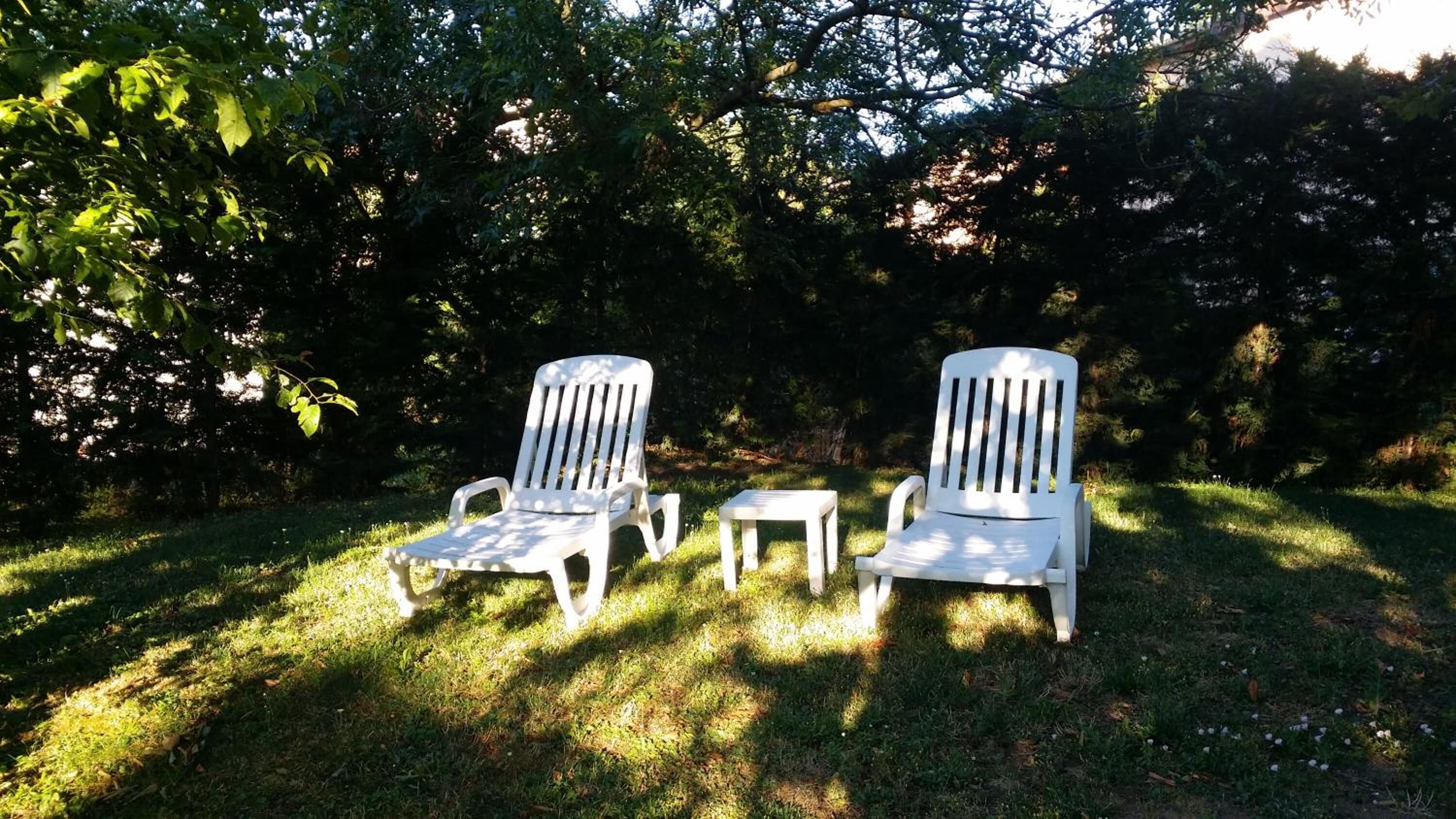 Two white Adirondack chairs and a small table in a grassy outdoor area with trees in the background.