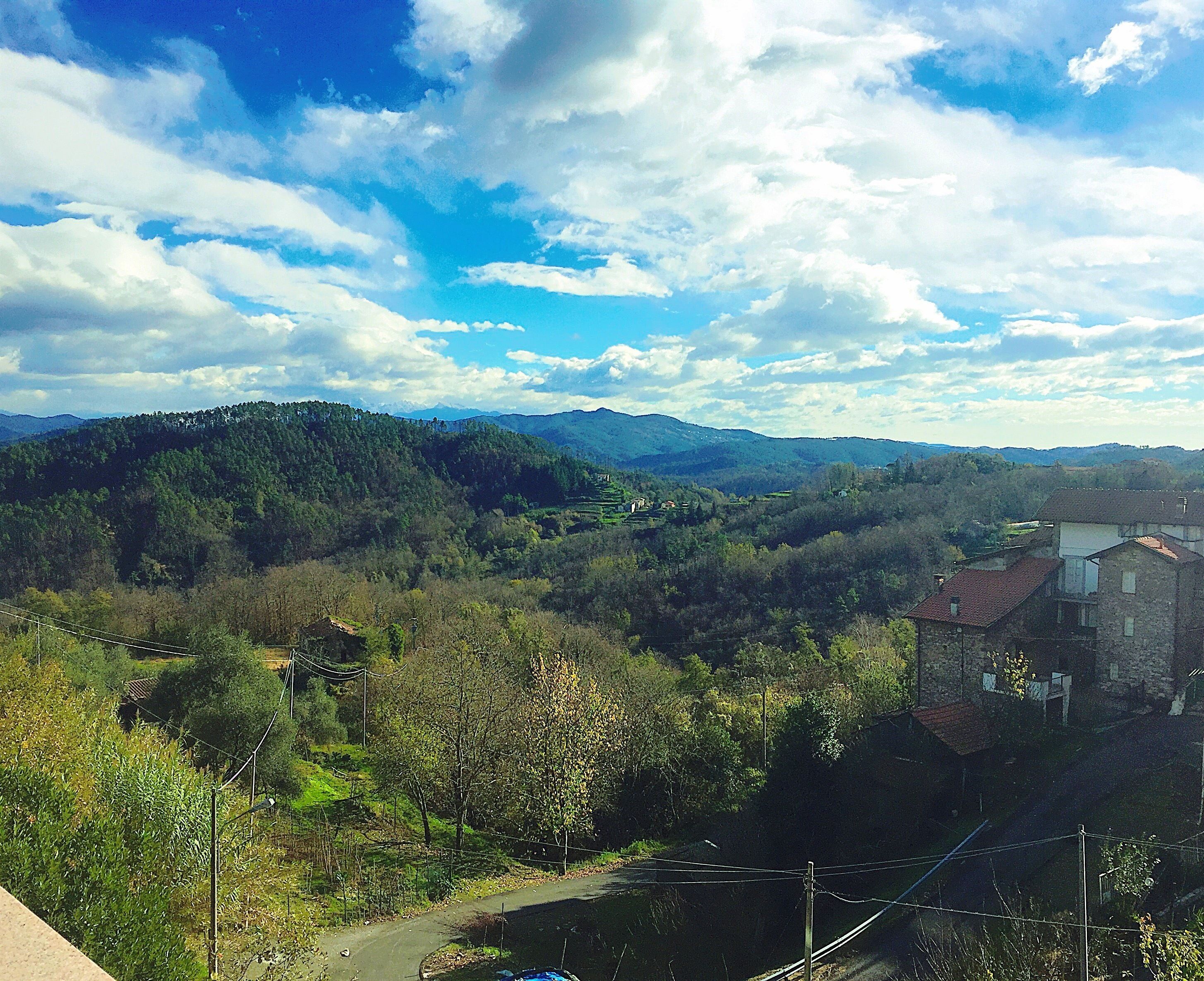 Un paysage rural avec un bâtiment en pierre, entouré d’une végétation luxuriante et des montagnes à l’horizon.