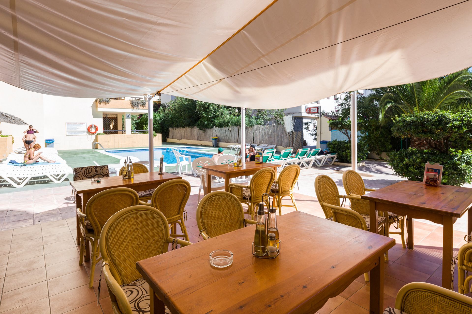 Un área de comedor al aire libre con sillas y mesas de mimbre, una piscina al fondo y una sección cubierta con sombrillas.