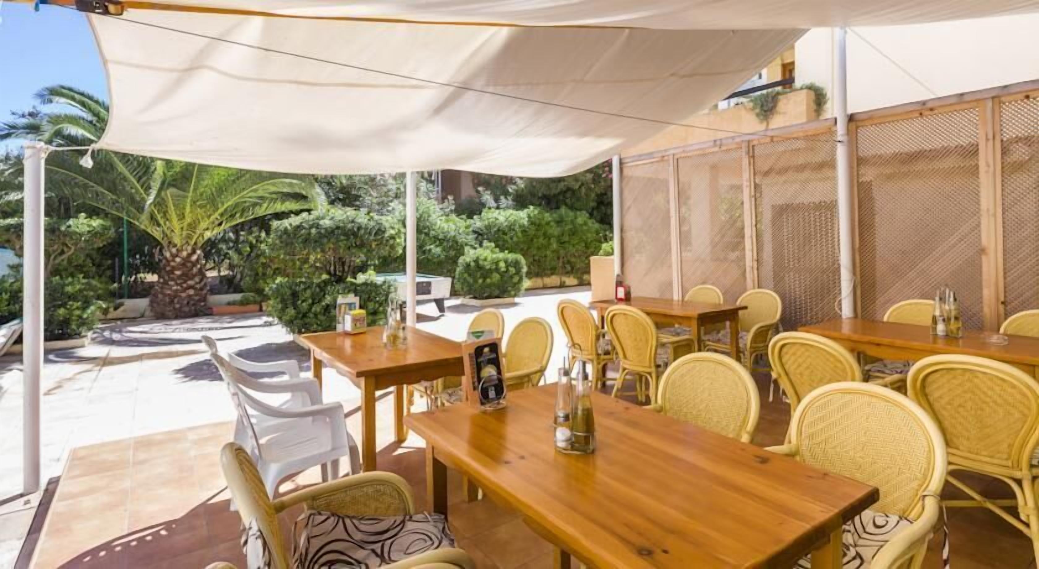 An outdoor dining area with wooden tables and wicker chairs, surrounded by a white canopy and a view of tropical plants.