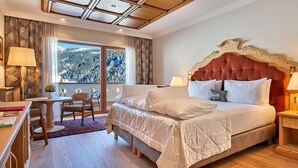 A hotel room with a large bed, a desk with a chair, a window with a view of a forest, and a ceiling with a wooden design. - Hotel Sassongher – Preferred Hotels & Resorts (Corvara in Badia)