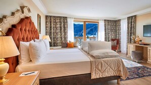 A hotel room with a large bed, a desk with a chair, a television, and a view of mountains through the window. - Hotel Sassongher – Preferred Hotels & Resorts (Corvara in Badia)