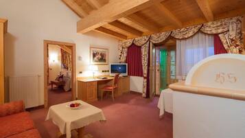 A cozy room with a wooden ceiling, a bed with a number, a desk with a computer, a table with fruit, and a view of a green-lit room through a window.