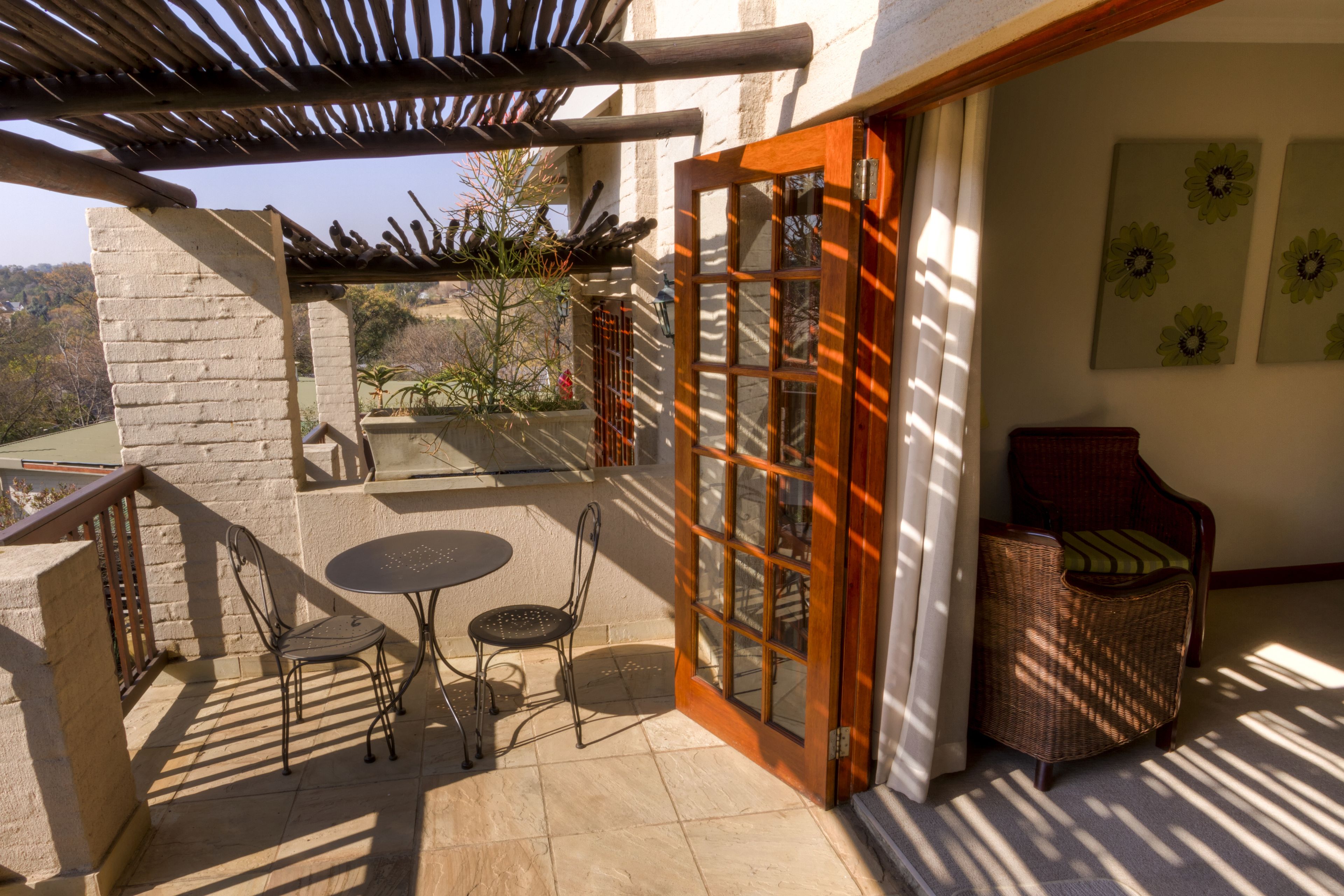 Une terrasse ensoleillée avec une table et des chaises, un fauteuil en osier et une porte en bois donnant sur un espace extérieur.