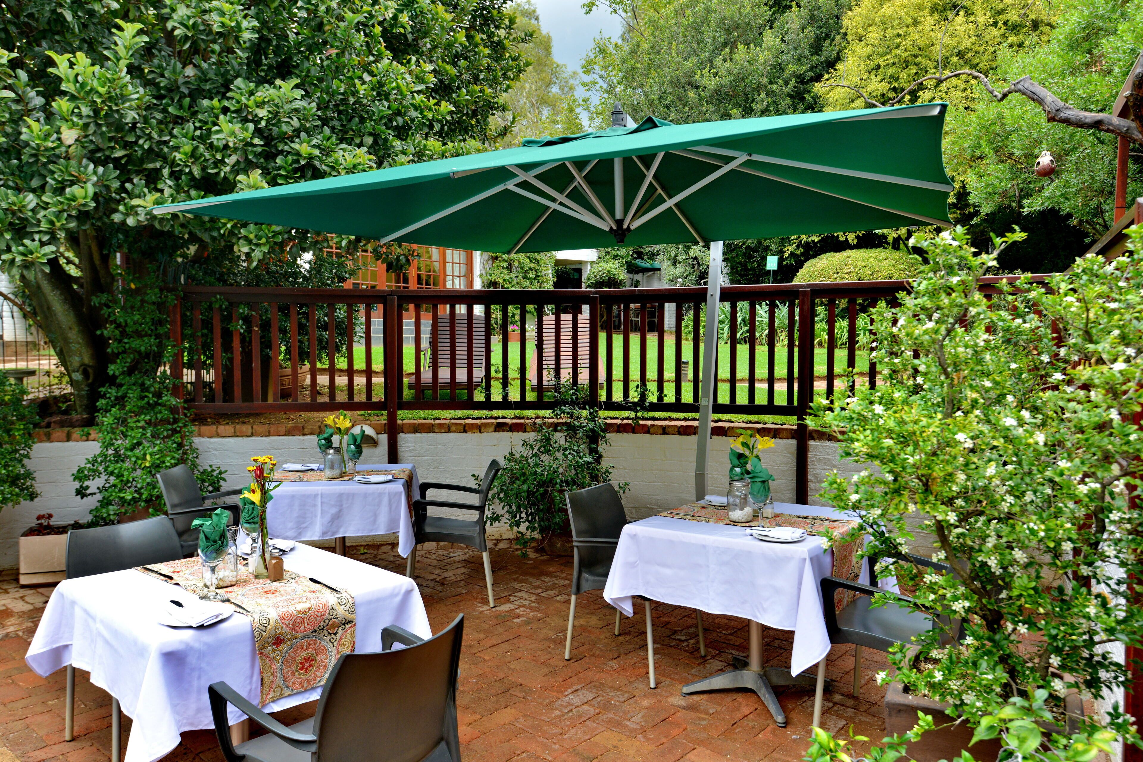 An outdoor patio with tables and chairs, covered by a green umbrella, surrounded by greenery and a brick wall.