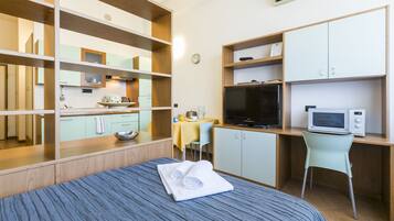 Una habitación de hotel con cama, escritorio con computadora, televisor y kitchenette.
