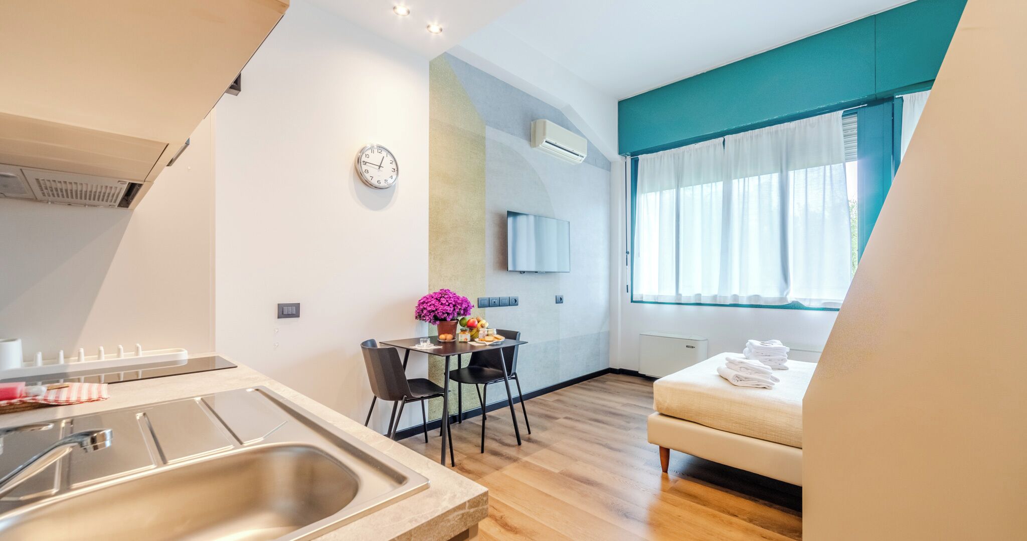A modern hotel room with a kitchen area, a dining table with chairs, a sofa, a TV mounted on the wall, and a window with curtains.