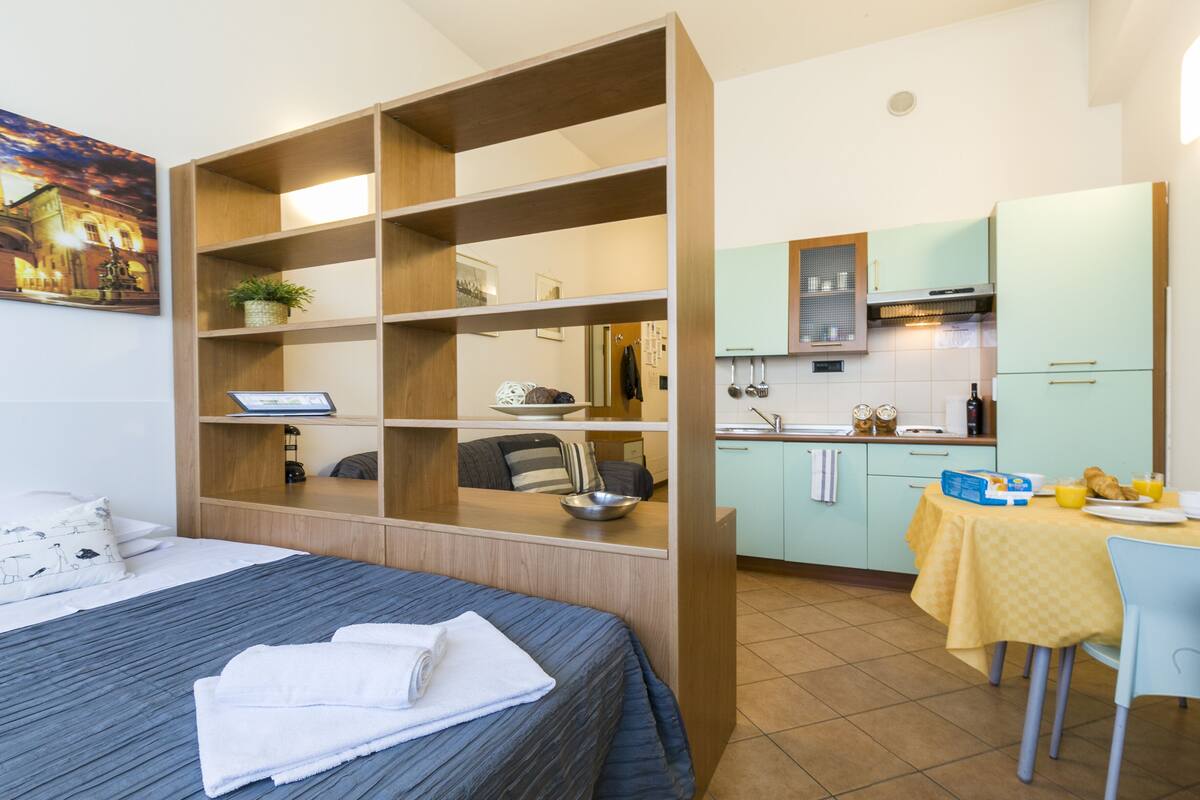 A compact hotel room with a bed, a built-in desk, a small kitchenette, and a dining area.