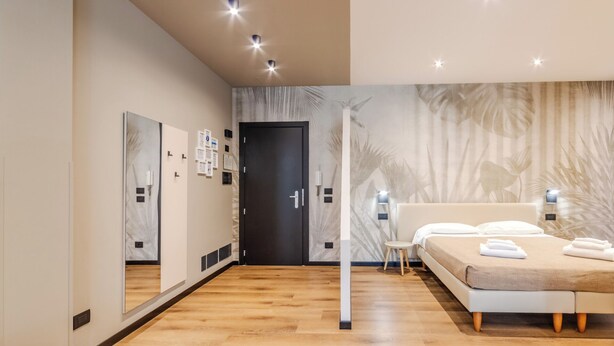 A modern hotel room with a wooden floor, a bed with white linens, a large mirror, and a wall with a tropical leaf pattern.