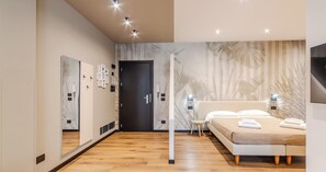 Habitación de hotel moderna con suelo de madera, cama con ropa blanca, un espejo grande y una pared con diseño de hojas tropicales.