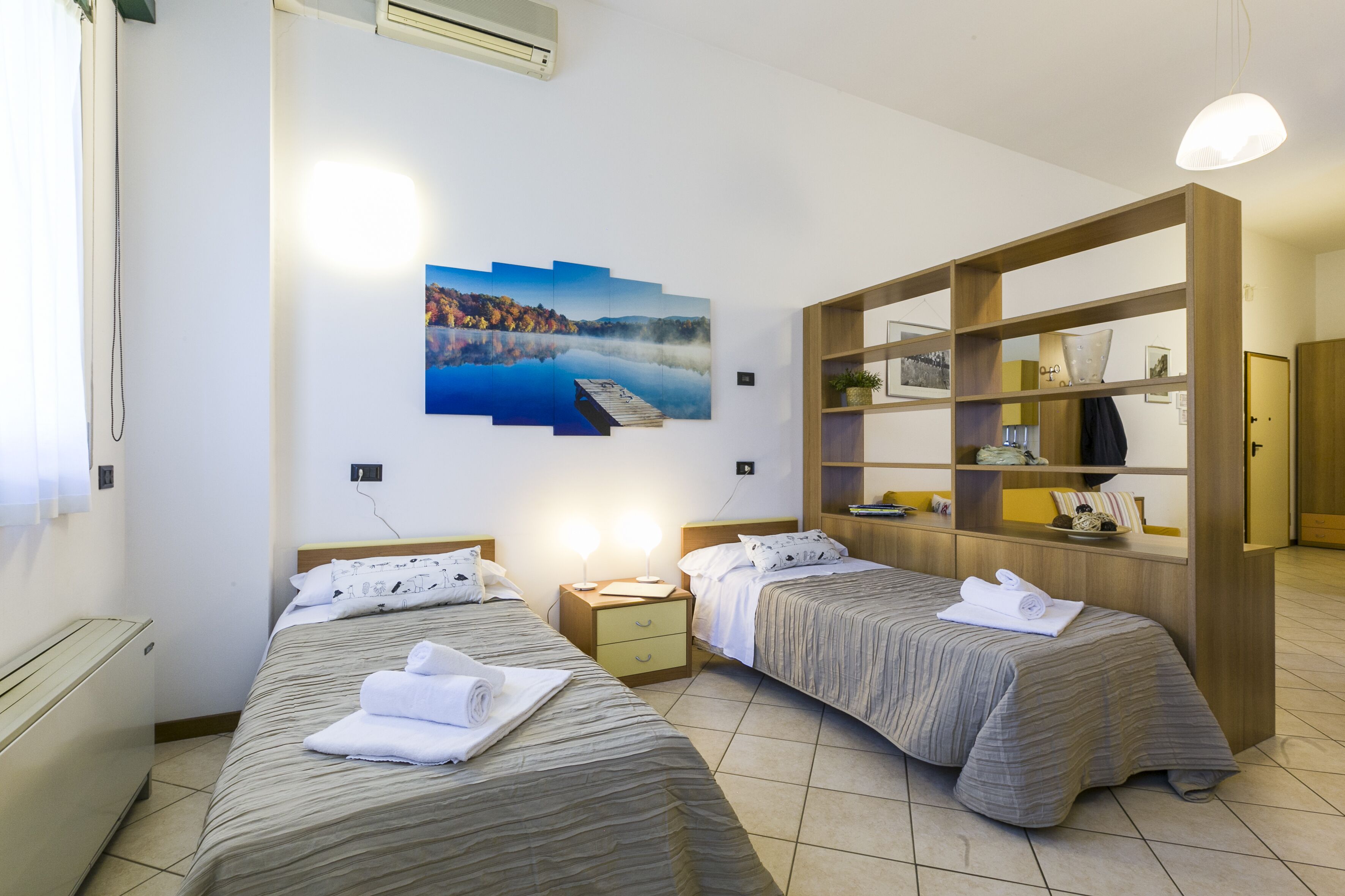 Une chambre d’hôtel avec deux lits, une bibliothèque en bois, un tableau représentant un lac et un petit bureau avec une lampe.