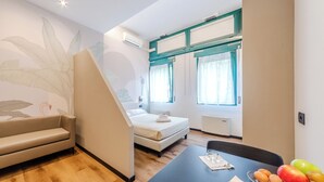 A compact room with a bed, a sofa, a dining table with fruit, and a wall with a leaf pattern.