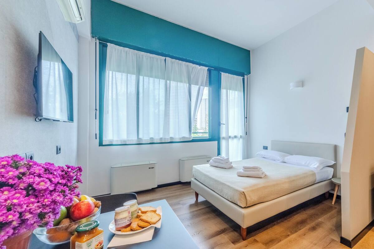 A hotel room with a bed, a sofa, a TV, and a table with food and flowers.