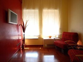 Una habitación con pared roja, ventana con cortinas traslúcidas, una mesita, una silla y una planta.