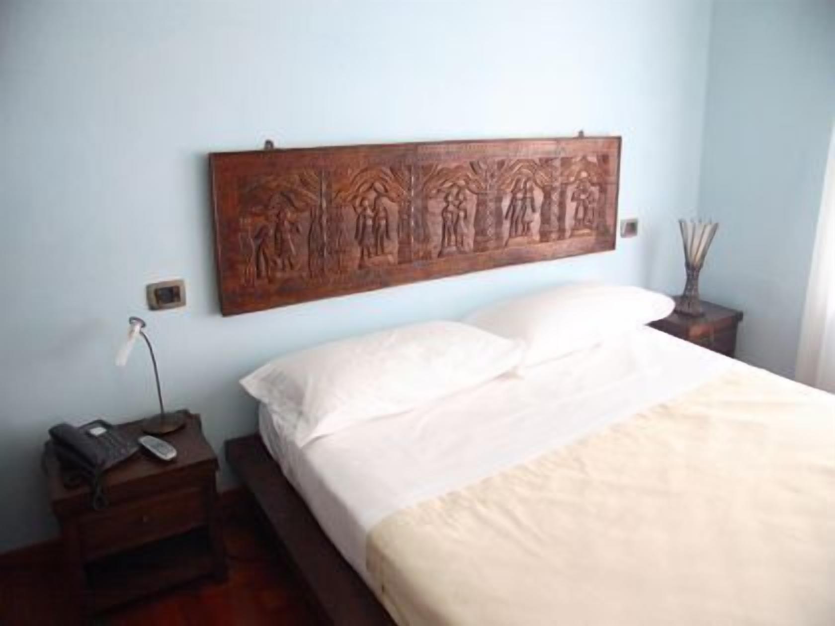 Un dormitorio con cabecera de madera, cama con ropa de cama blanca, mesita de noche con lámpara y un pequeño adorno en la mesita.