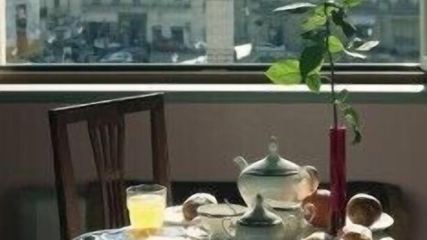 Une table avec une théière, des tasses et une rose dans un vase, une chaise, et une fenêtre donnant sur une statue et des bâtiments en arrière-plan.