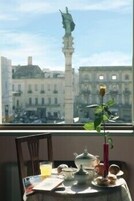 Uma mesa com um bule, xícaras e uma rosa em um vaso, uma cadeira e uma janela com uma estátua e edifícios ao fundo.