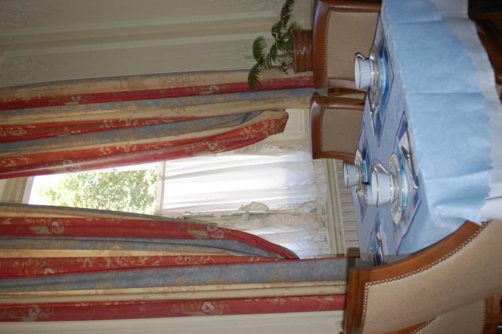 A room with a large window, a patterned curtain, and a wooden table with a blue tablecloth.