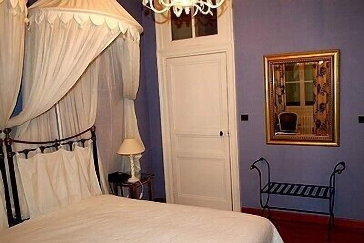A bedroom with a canopy bed, a door, a small table with a lamp, and a chair.