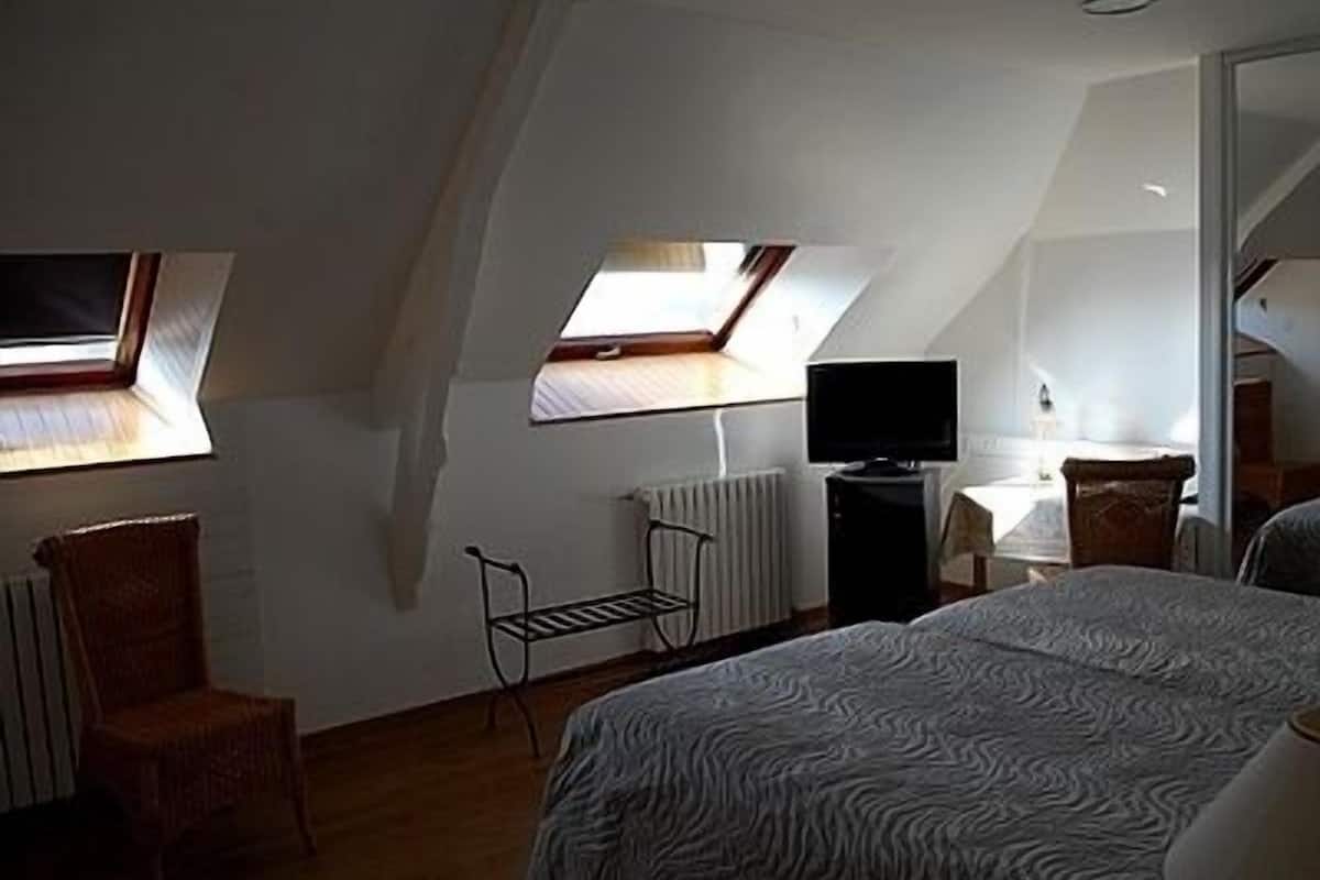 A bedroom with a sloped ceiling, a bed, a chair, a desk with a computer, and a television.