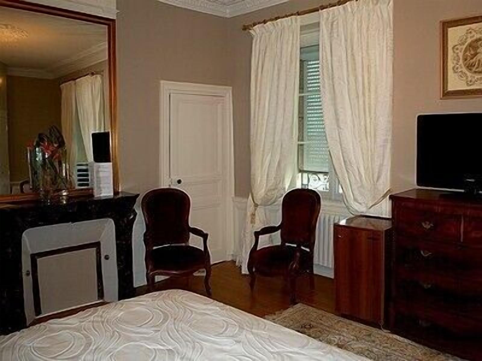 Une chambre à coucher comprenant un lit, deux chaises, une commode, un téléviseur et une fenêtre avec des rideaux.