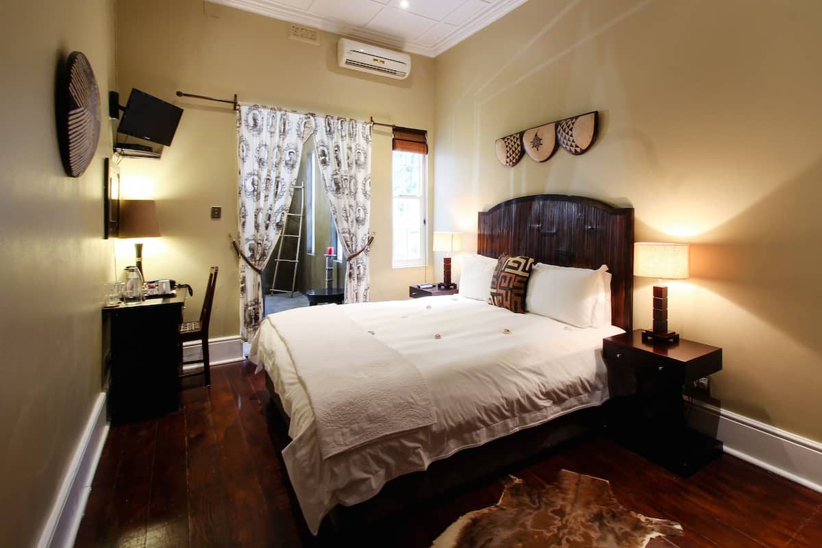 A hotel room with a large bed, a wooden headboard, a small table with a lamp, a wall-mounted TV, and a window with curtains.