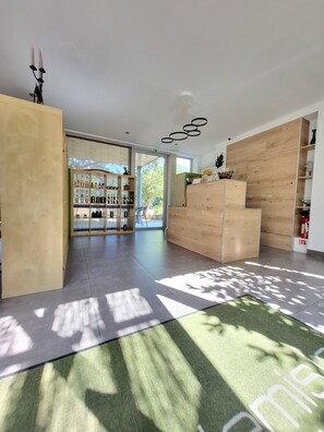 A modern hotel lobby with a reception desk, a bar area with shelves of bottles, and a view of greenery outside.