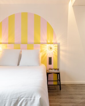 A bedroom with a bed, a bedside table with books, and a wall with a striped pattern. - DEMAIN Hôtel & Conciergerie (Nantes)