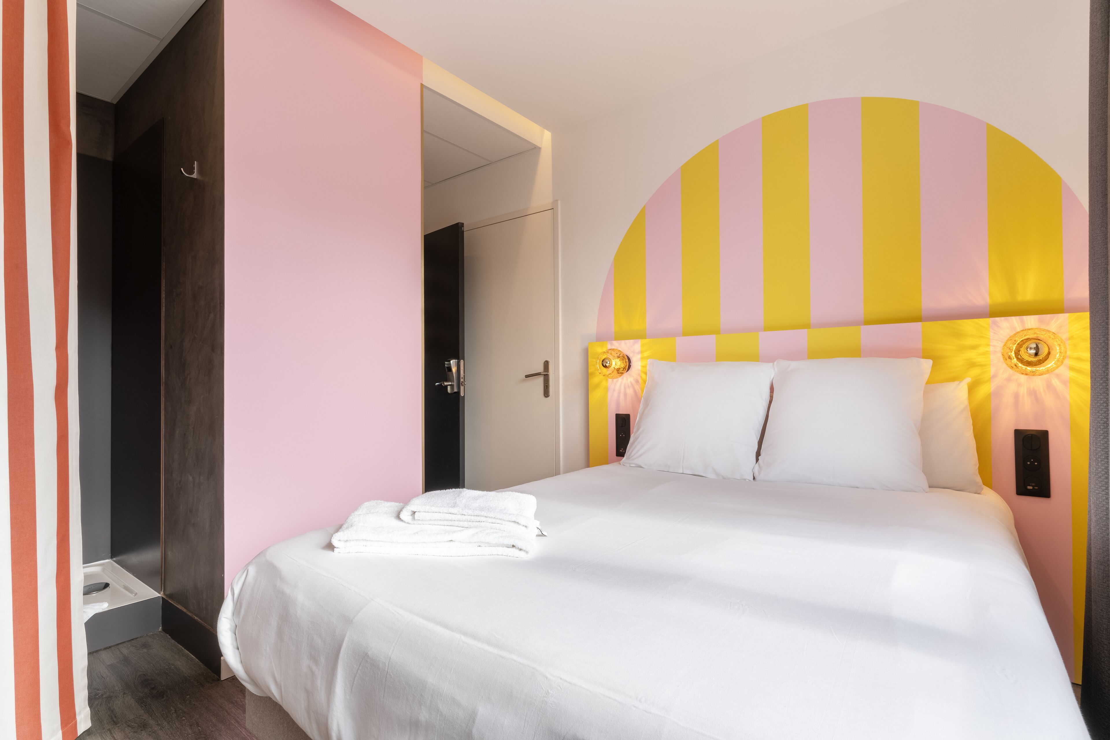 A neatly made bed with white linens and a folded towel, a headboard with a yellow and pink striped design, and a door leading to a bathroom.