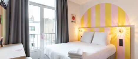 Habitación de hotel con cama, escritorio, silla, ventana con cortinas y televisor montado en la pared.