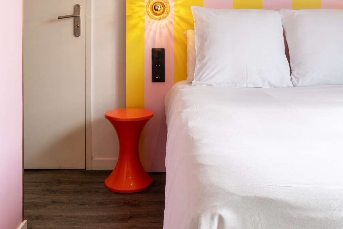 A bedroom with a bed, a red stool, a door, and a wall with a striped pattern.