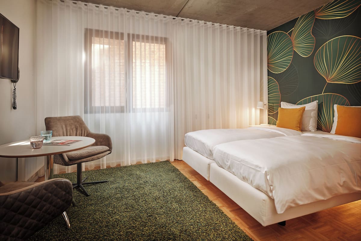 A hotel room with a bed, a chair, a small table, and a window with sheer curtains.