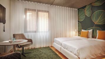 Una habitación de hotel con cama, una silla, una mesita y una ventana con cortinas traslúcidas.