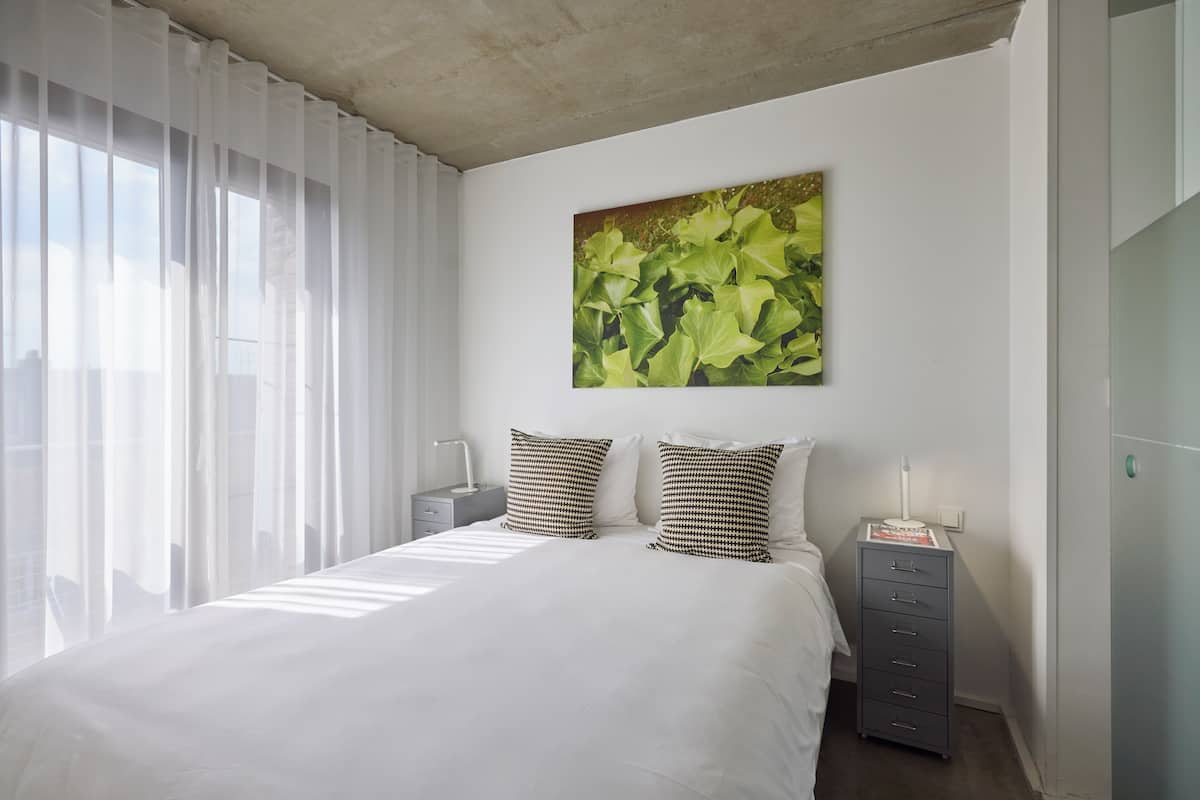 A bedroom with a large bed, white curtains, a wall-mounted artwork, and a bedside table with a lamp.