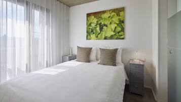 Un dormitorio con una cama grande, cortinas blancas, un cuadro en la pared y una mesita de noche con una lámpara.