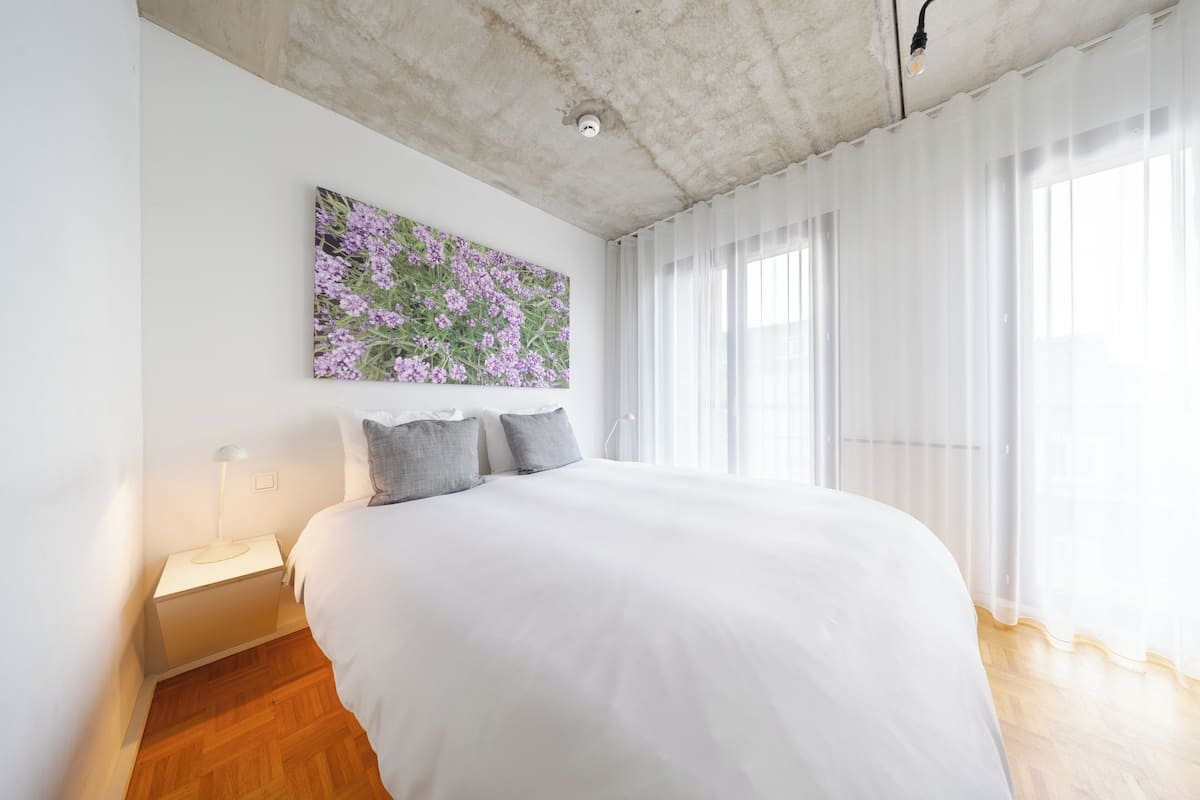 A modern bedroom with a large bed, a nightstand, a wall-mounted lamp, and a floral artwork.