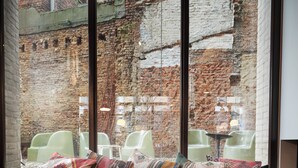 Een moderne woonkamer met een zwarte bank, kleurrijke sierkussens, een klein rond tafeltje en uitzicht op een stadsstraat door grote ramen.