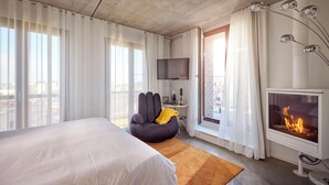 Een moderne hotelkamer met een groot bed, een comfortabele fauteuil, een flatscreen televisie, een open haard en een balkon met uitzicht op de stad.