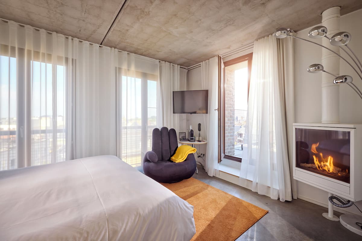A modern hotel room with a large bed, a comfortable armchair, a flat-screen TV, a fireplace, and a balcony with a city view.
