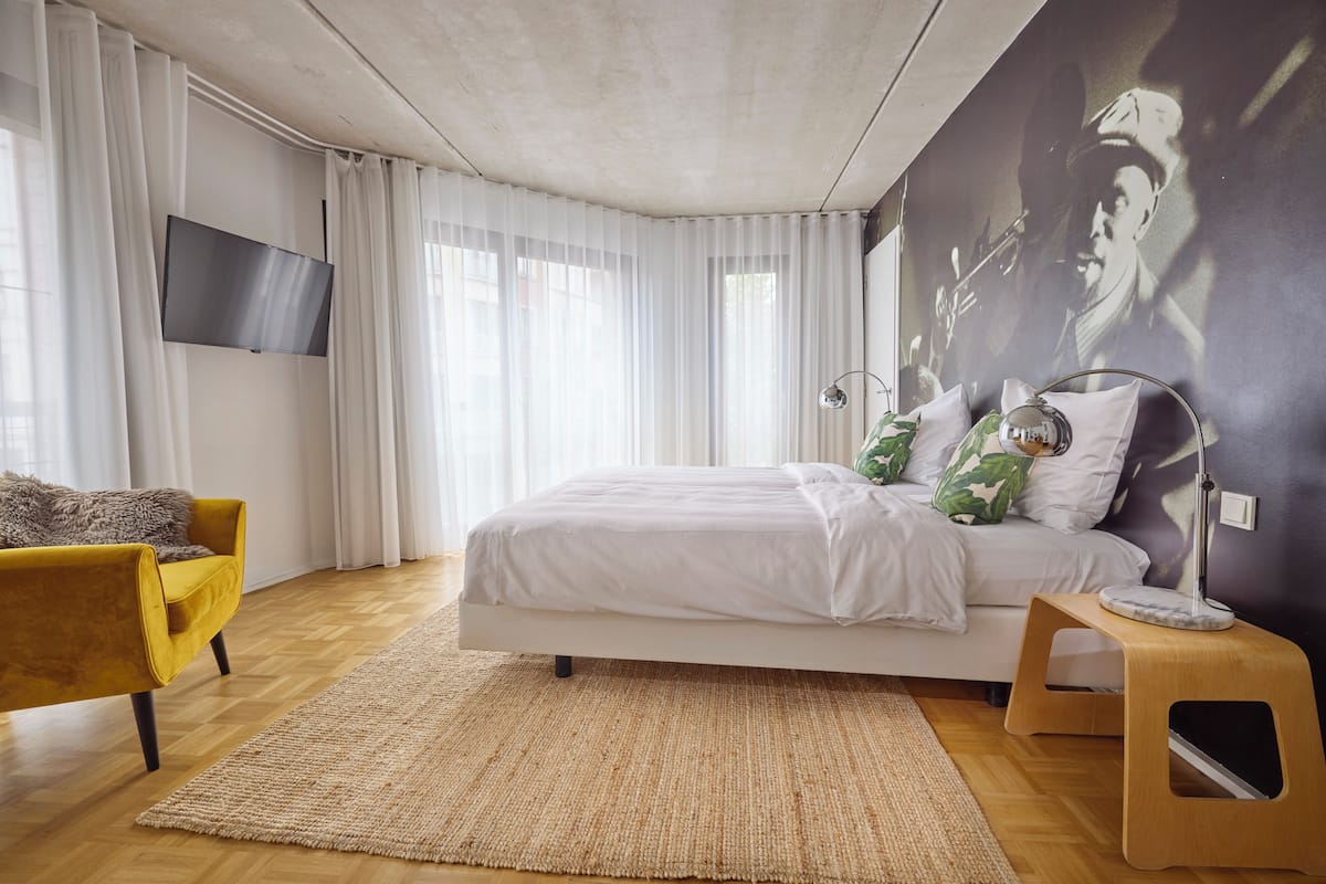 A modern bedroom with a bed, a yellow armchair, a small wooden stool, and a wall-mounted TV.