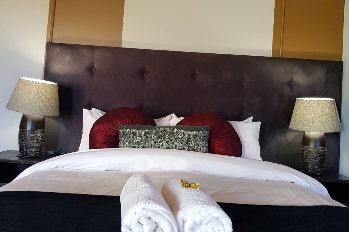 Una cama bien hecha con sábanas blancas, dos almohadas rojas y una almohada decorativa con un estampado floral verde. Hay dos lámparas de noche con pantallas beige a cada lado de la cama.