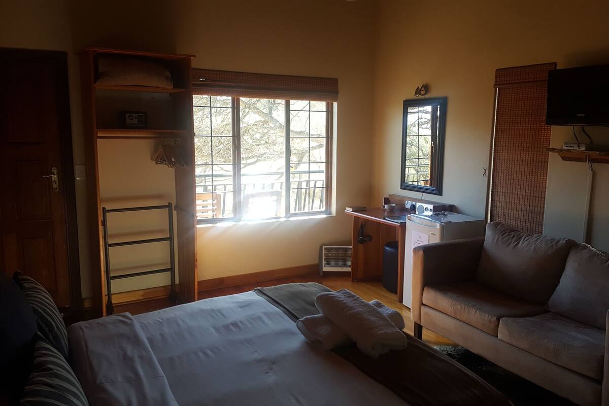 Un dormitorio con cama, escritorio, silla, televisor y ventana con vista a árboles.
