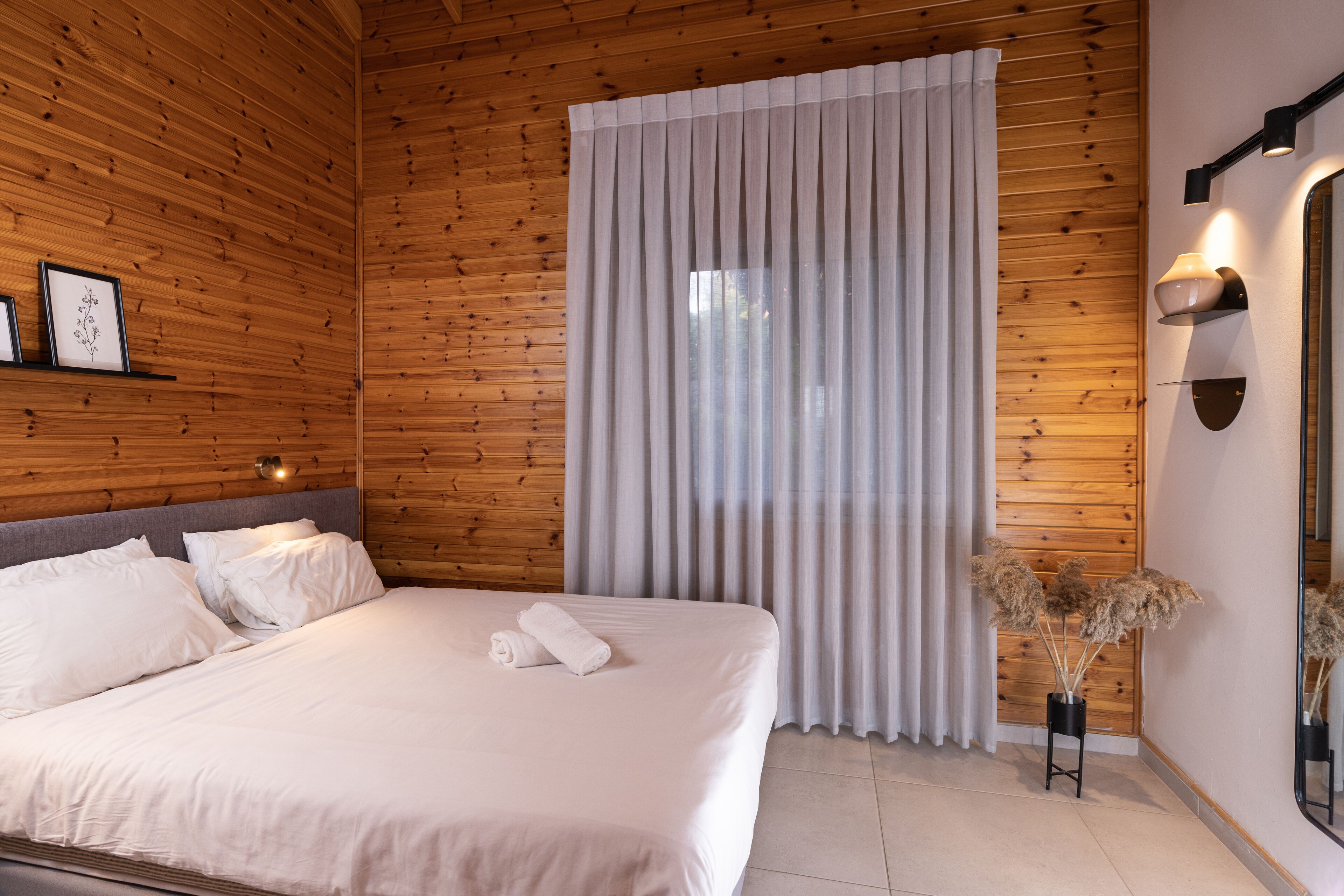 Un lit bien fait, avec du linge de lit blanc et une serviette pliée, une tête de lit en bois, une grande fenêtre avec des rideaux légers, un luminaire fixé au mur et un miroir sur le côté droit.