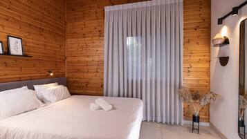 Un lit bien fait, avec du linge de lit blanc et une serviette pliée, une tête de lit en bois, une grande fenêtre avec des rideaux légers, un luminaire fixé au mur et un miroir sur le côté droit.