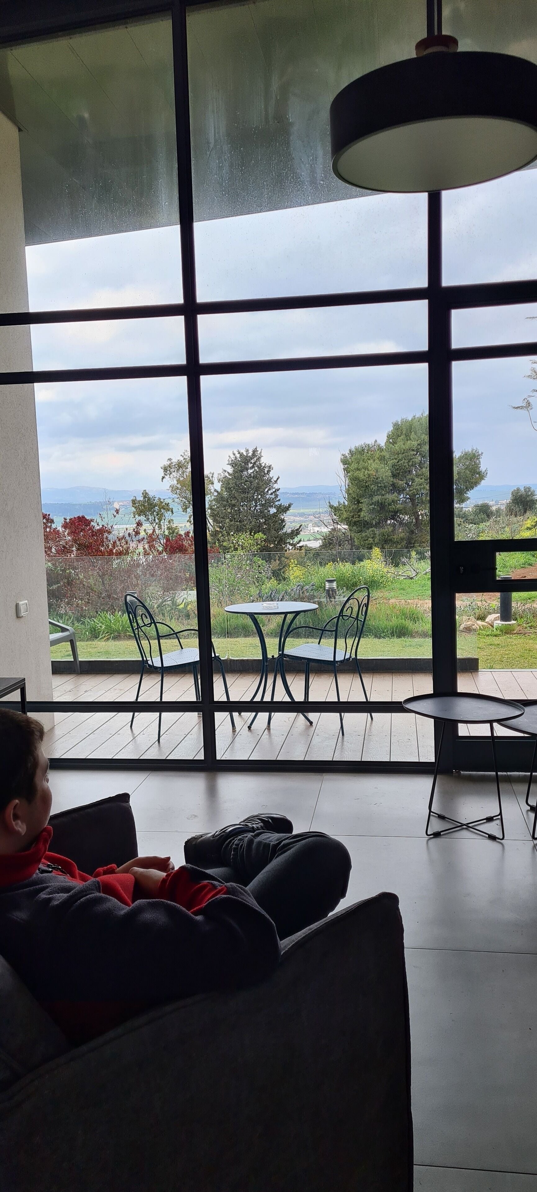 Quarto com grandes janelas dando para um jardim e uma vista distante. Uma criança está sentada num sofá, e há uma mesa com cadeiras do lado de fora.